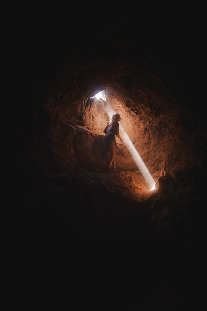 photo of woman standing inside a cave in a beam of light that is streaming in from opening