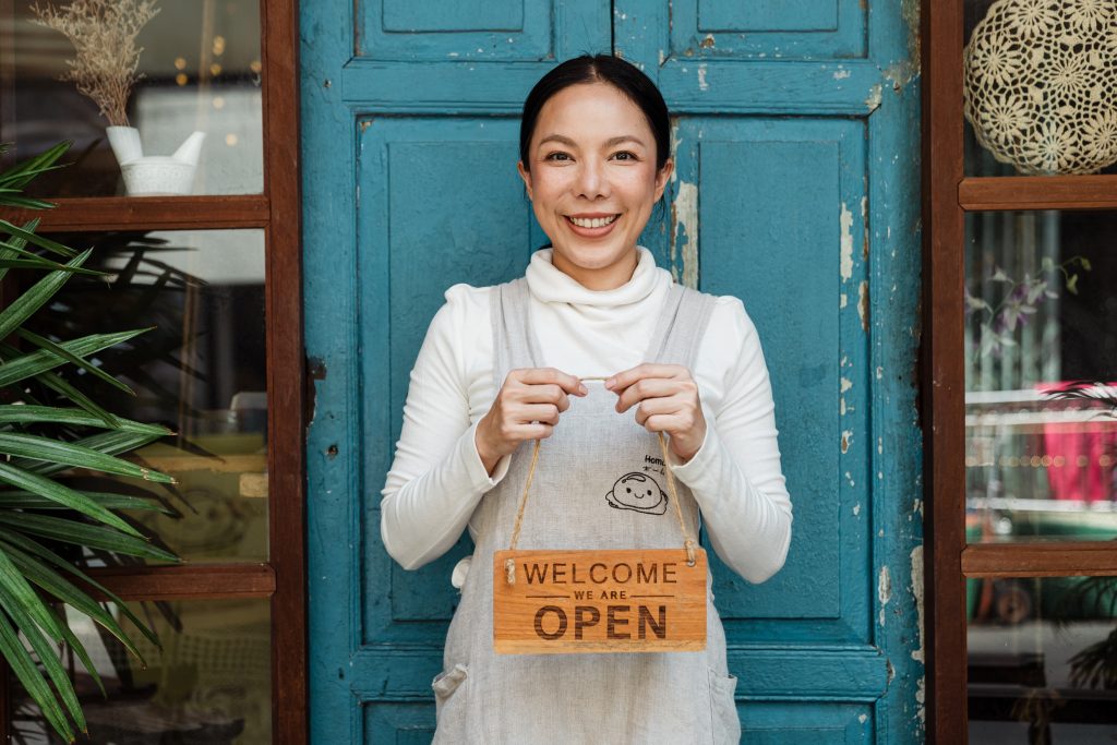 woman entrepreneur standing outside door holding "welcome - we are open" sign